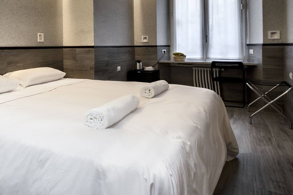 A modern hotel room with a large bed, a bedside table with a lamp, a window with curtains, and a radiator.