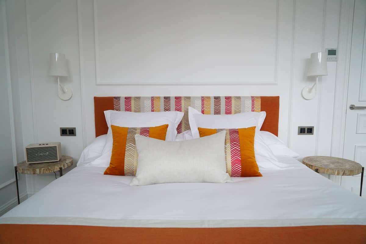 Ein ordentlich bezogenes Bett mit weißer und orangefarbener Bettwäsche, zwei Nachttische und eine Wandlampe.
