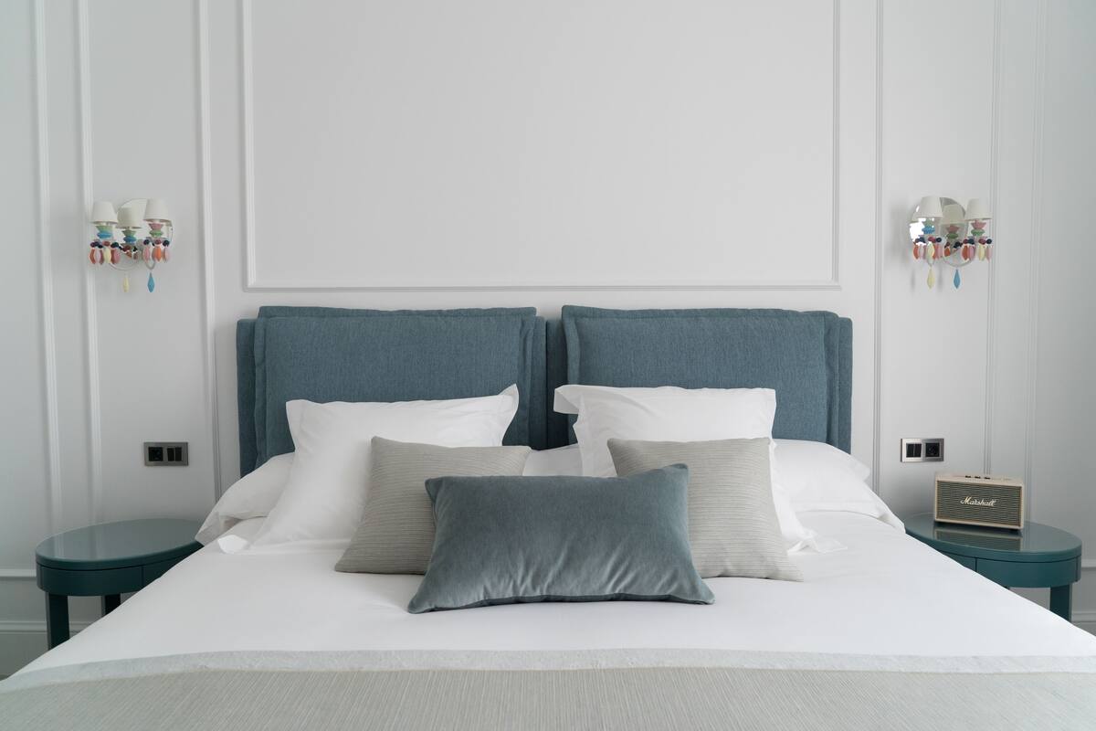 Ein ordentlich bezogenes Bett mit blauem Kopfteil, weißen Kissen und einer grauen Decke. Auf beiden Seiten des Bettes befinden sich Nachttische, auf dem rechten Tisch steht ein Radio.
