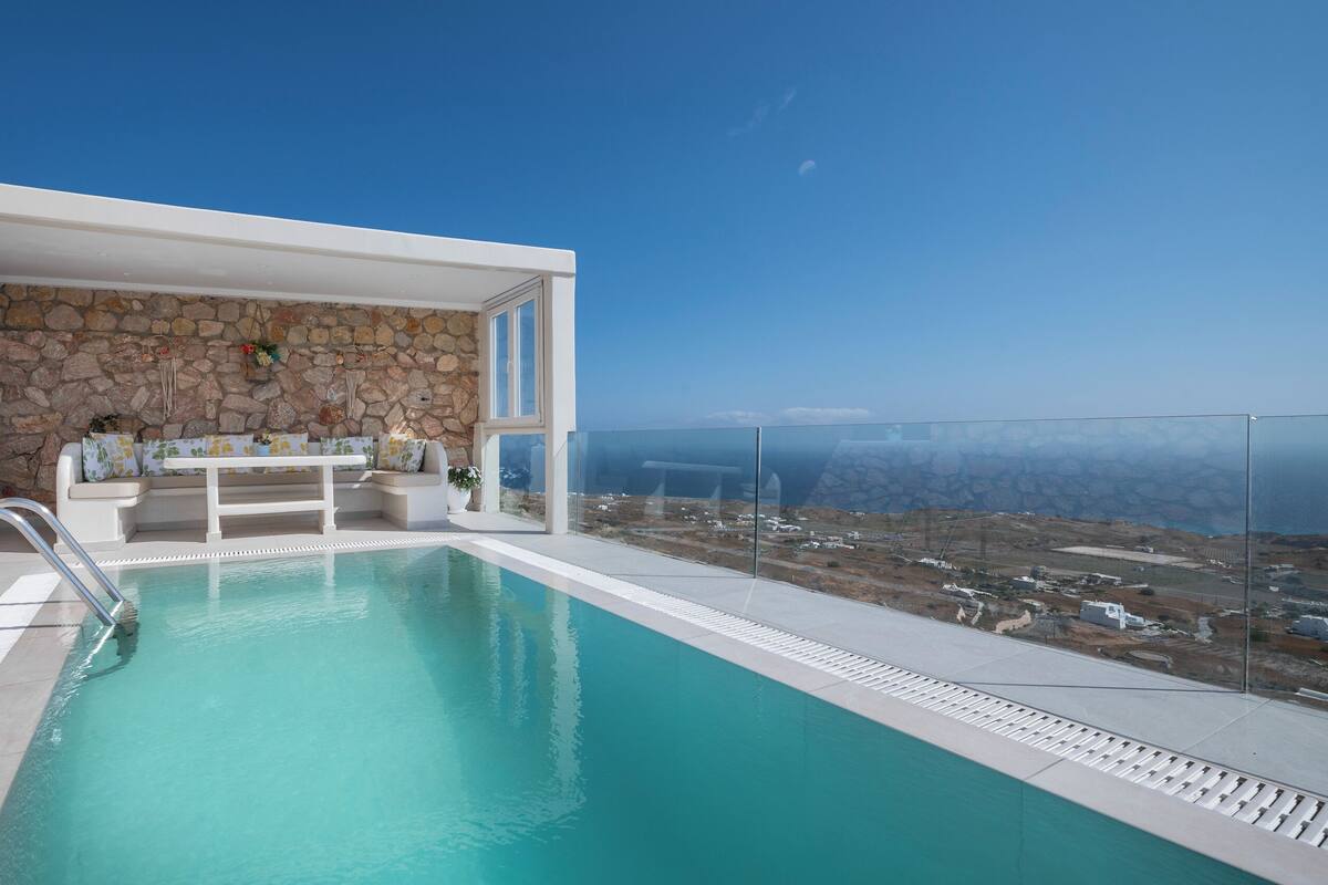 A modern outdoor pool area with a stone wall backdrop, a seating area with cushions, and a clear view of the landscape.