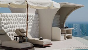 A poolside lounge area with a white umbrella, lounge chairs, and a small table. - Dream Villa (Santorini)