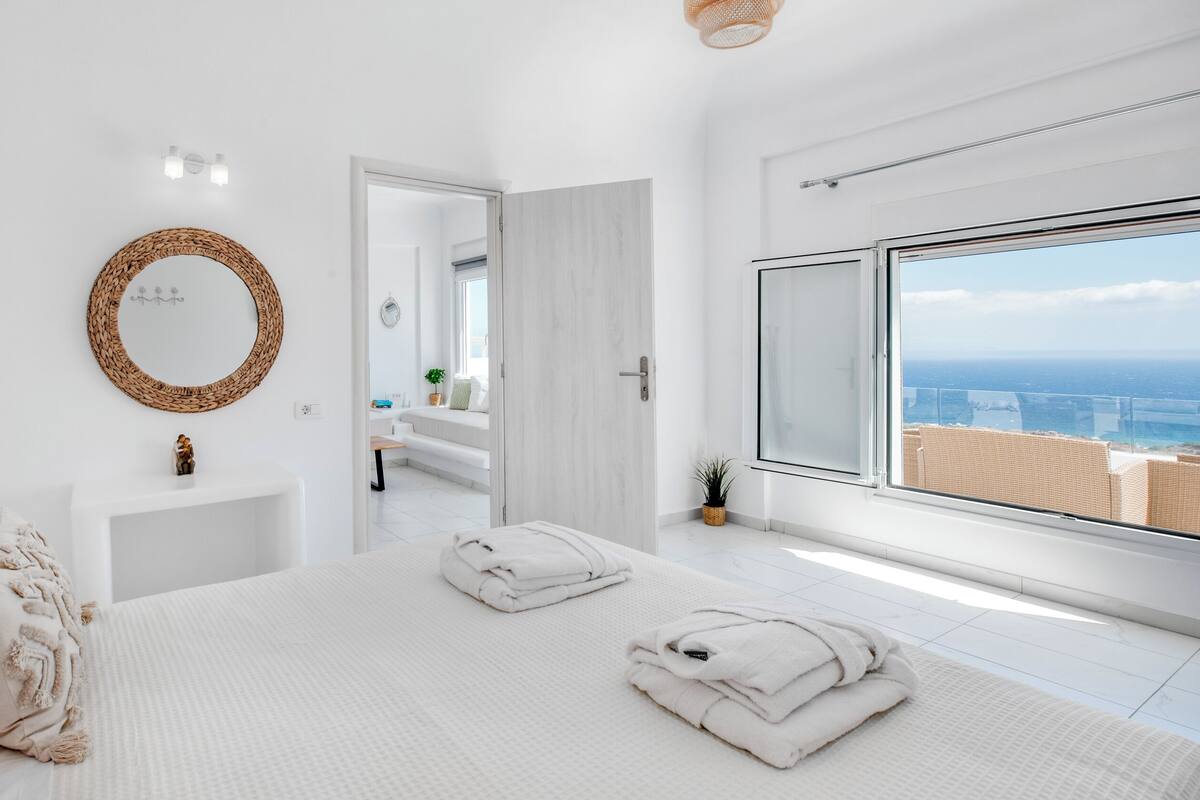 A modern bedroom with a large bed, a round mirror, a small bedside table, and a view of the ocean through a large window.