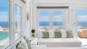 A modern, minimalist room with large windows offering ocean views, a comfortable seating area with patterned cushions, and a small potted plant. - Dream Villa (Santorini)