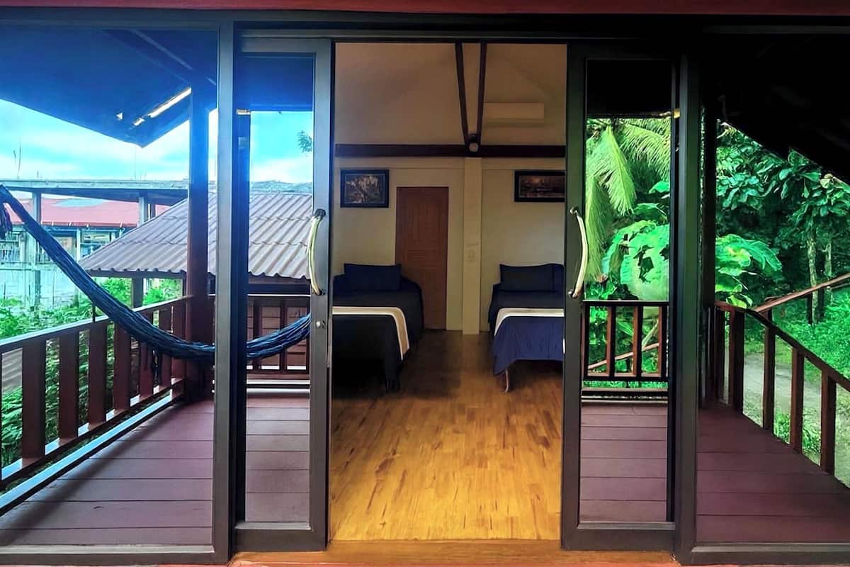 A room with two beds, a wooden floor, and a view of lush greenery through a glass door.