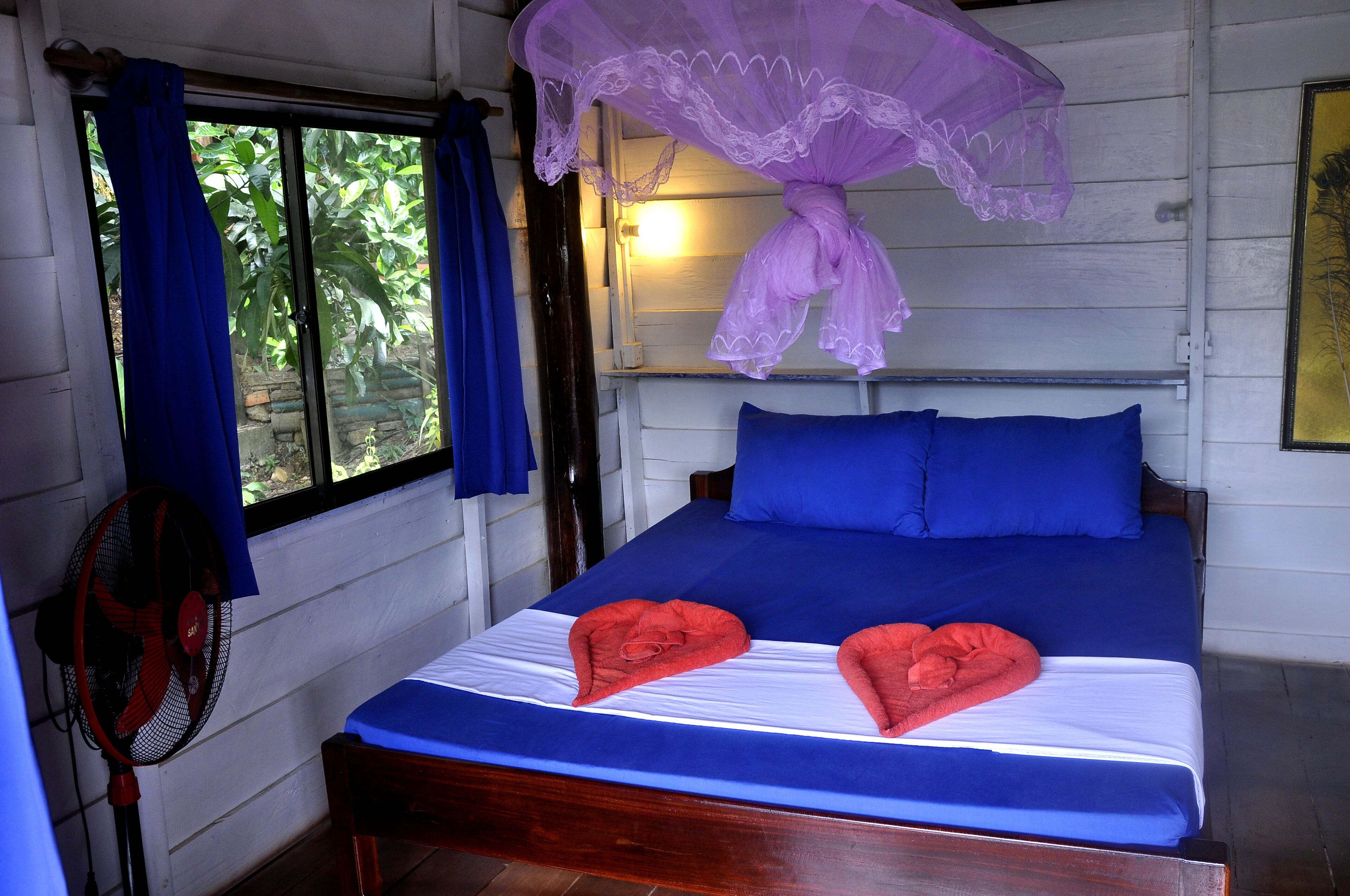 A bed with blue bedding and two red heart-shaped towels, a standing fan, and a window with blue curtains.