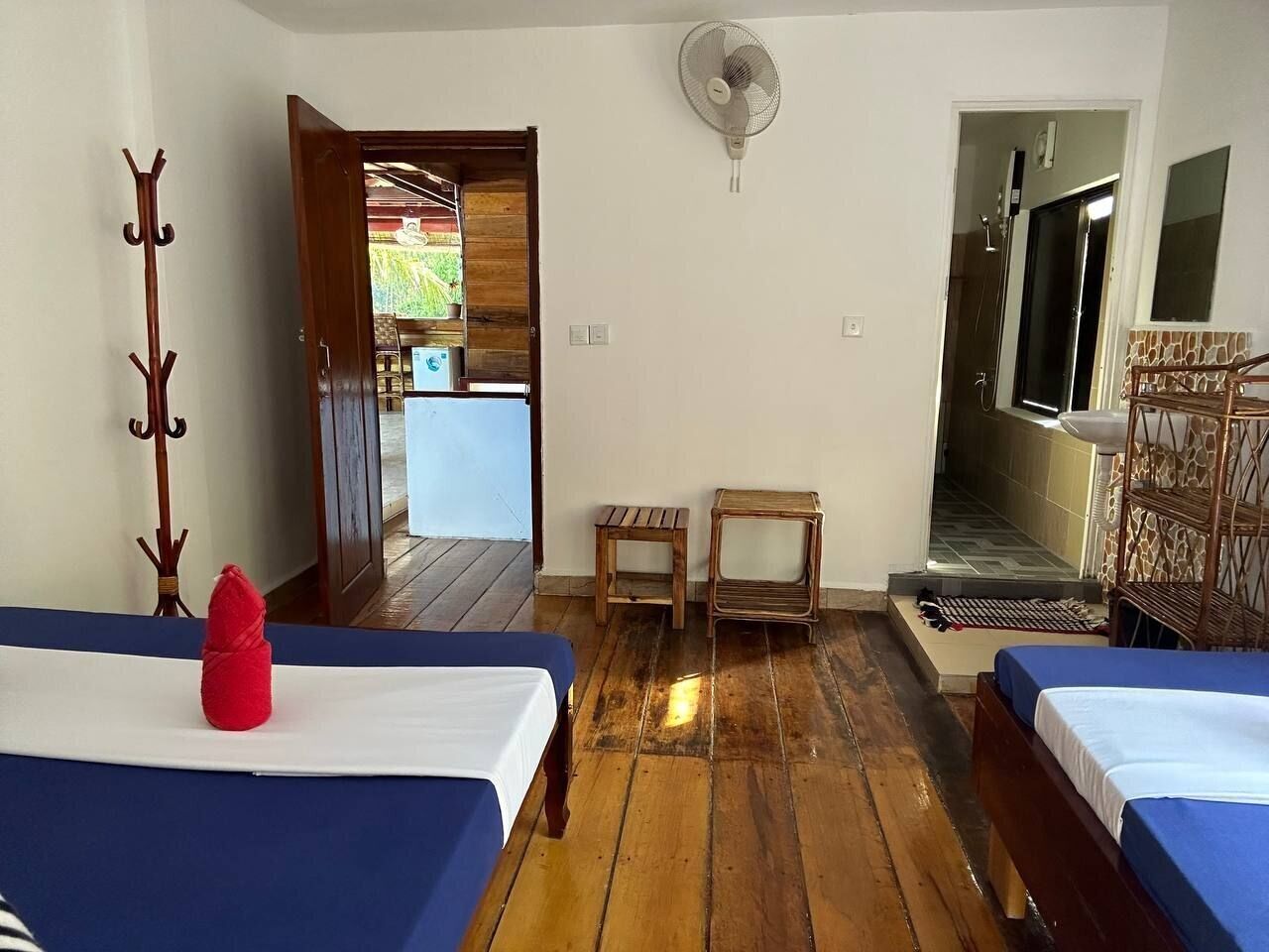 Une chambre avec deux lits, un plancher en bois, un ventilateur et une vue sur un jardin, accessible par une porte ouverte.