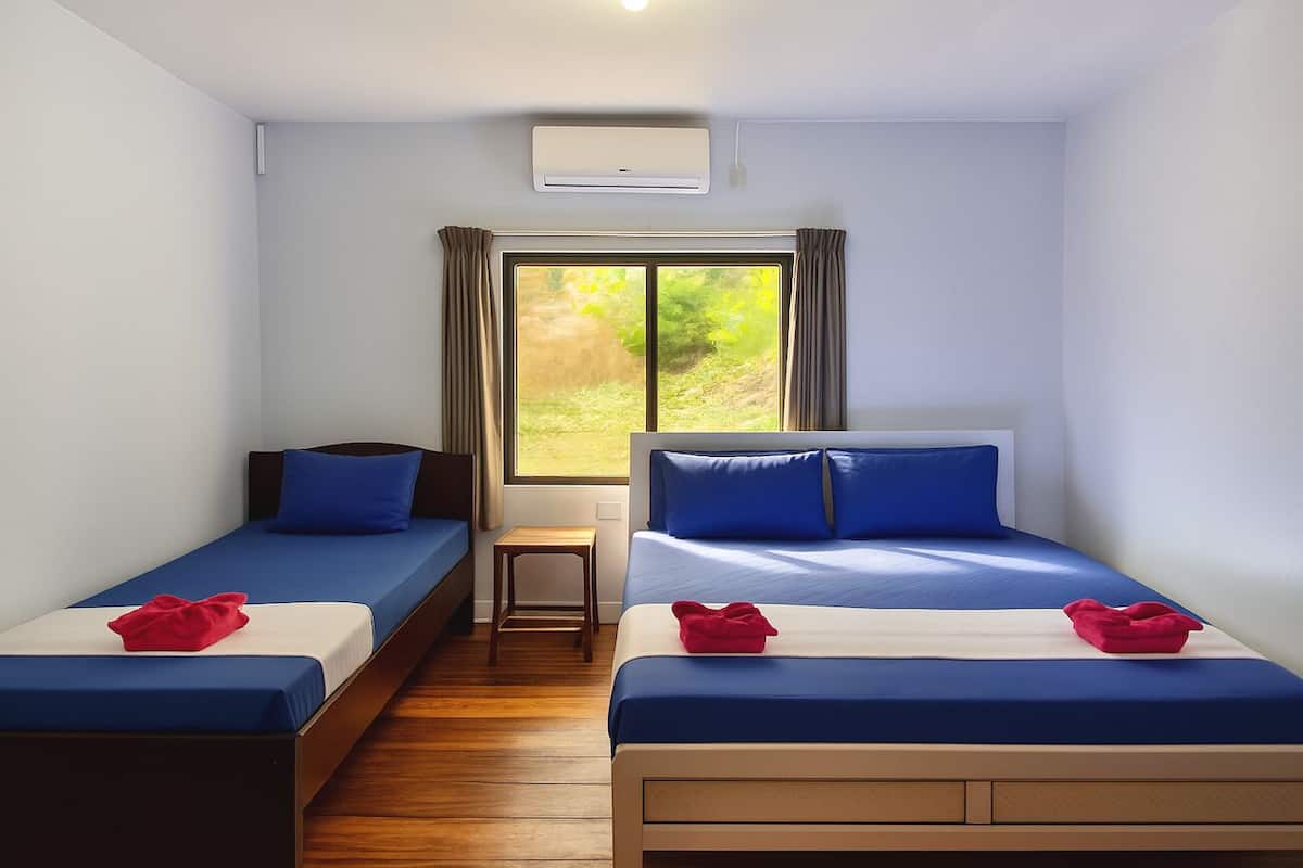 A room with two beds, a window with curtains, a small table, and an air conditioning unit.