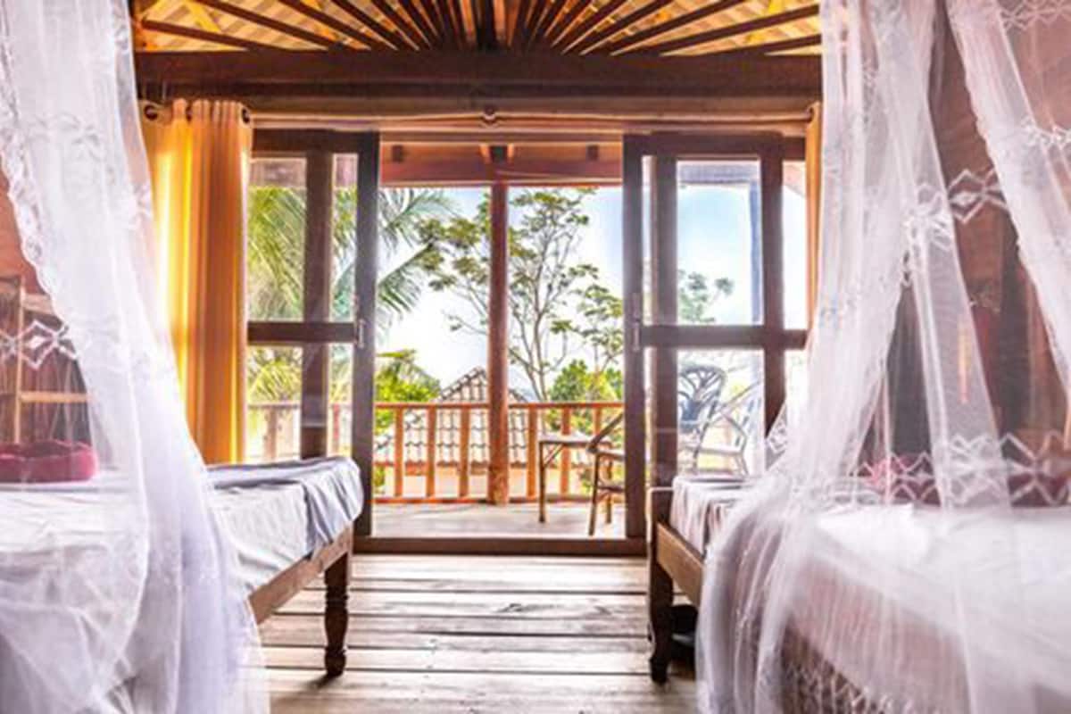 A room with two beds, a wooden ceiling, and large windows overlooking a balcony with a view of trees and a clear sky.