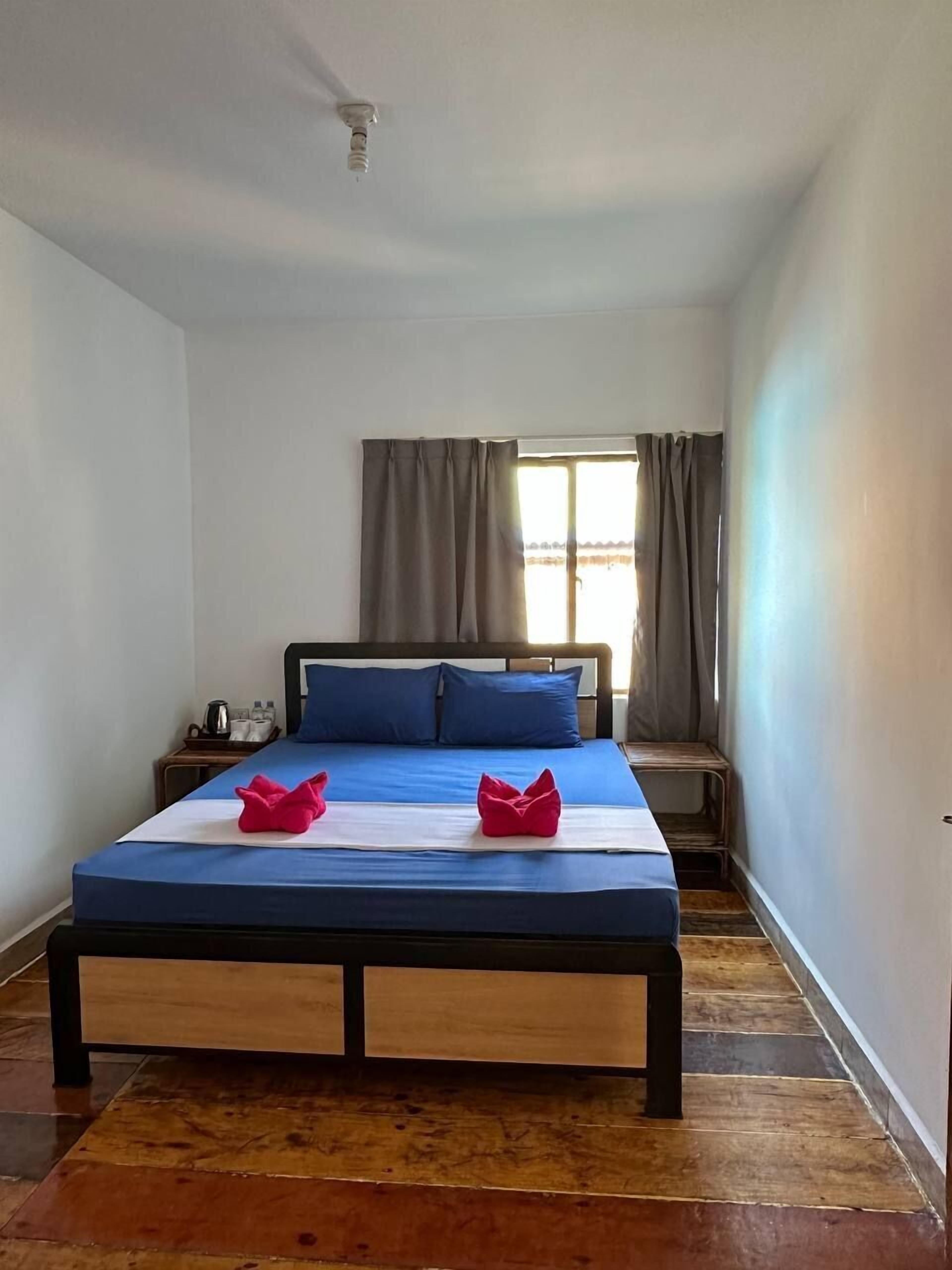 Un dormitorio con cama, almohadas, una mesita de noche, una ventana con cortinas y un ventilador de techo.