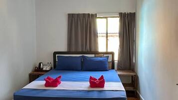 Un dormitorio con cama, almohadas, una mesita de noche, una ventana con cortinas y un ventilador de techo.