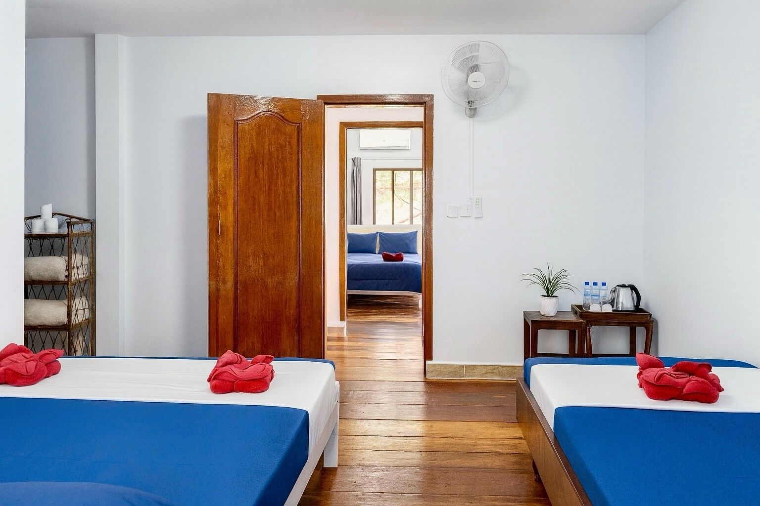 A hotel room with two beds, wooden flooring, a wooden wardrobe, a bedside table with a plant, and a view into another room with a bed and a blue blanket.