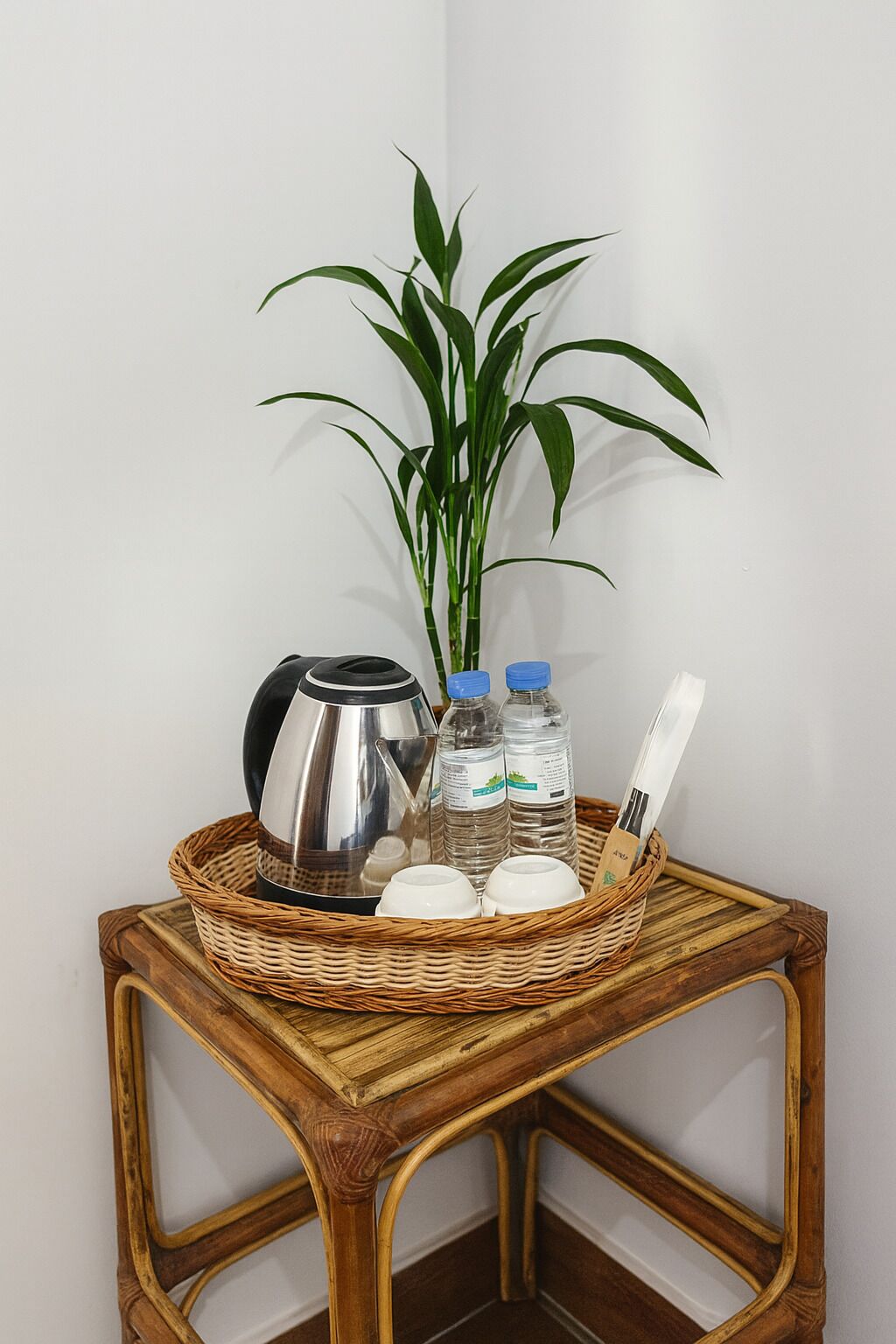 A corner table with a woven basket containing a kettle, water bottles, and cups, a bamboo plant, and a wooden spoon.