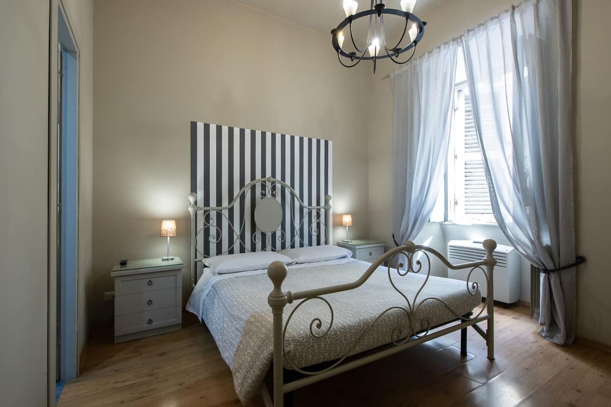 Quarto com cama, mesas de cabeceira, espelho, candelabro, cortinas e janela com persianas.