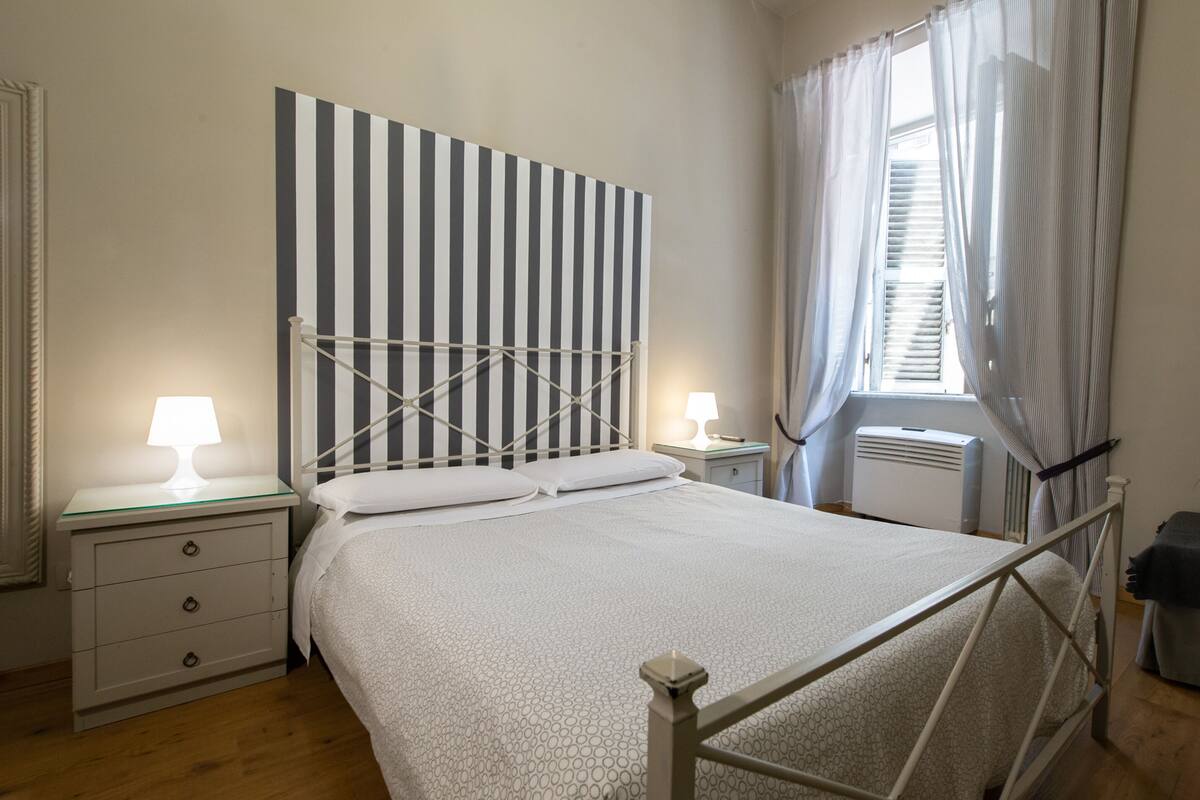 Quarto com cabeceira listrada, duas mesas de cabeceira com luminárias, cama com roupa de cama branca e uma janela com cortinas.