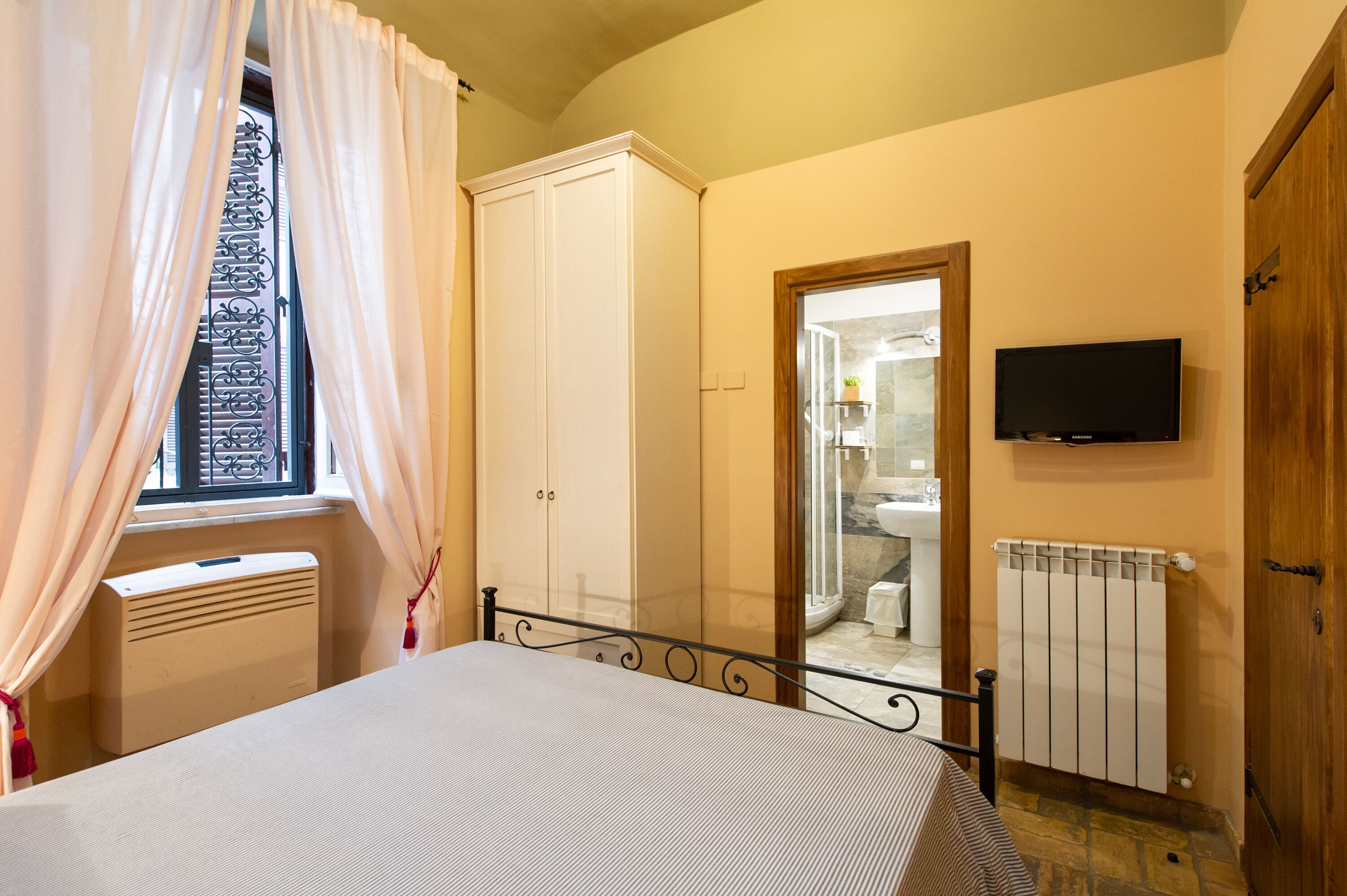 A bedroom with a bed, a radiator, a television, and a bathroom visible through an open door.