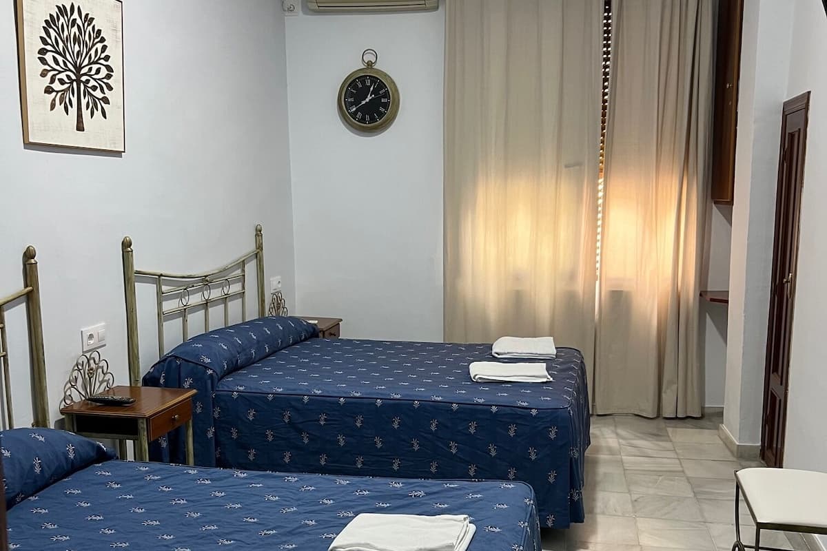 A hotel room with two beds, a clock, an air conditioner, and a window with curtains.