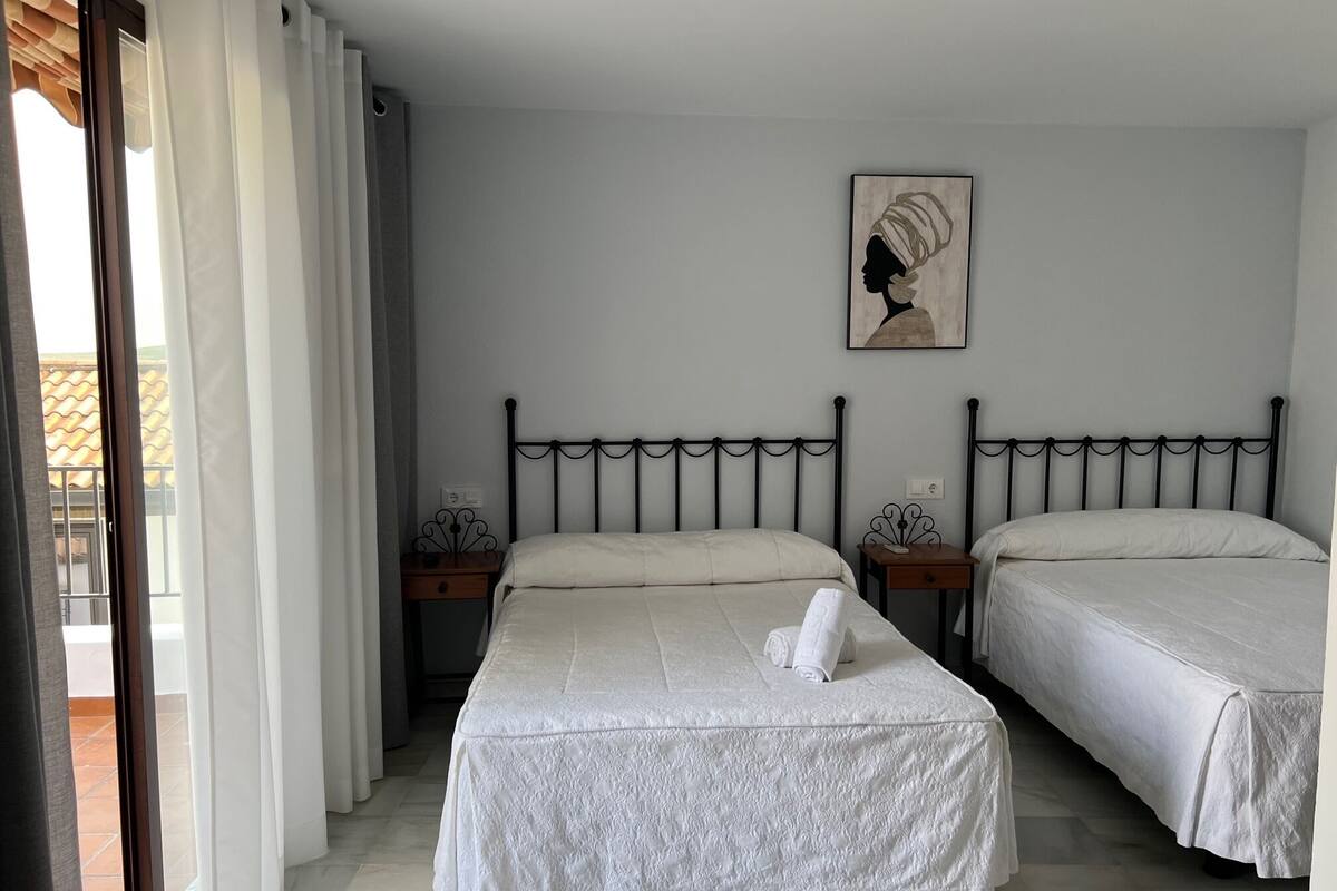 A bedroom with two beds, a window with curtains, a painting on the wall, and a small table with a lamp.