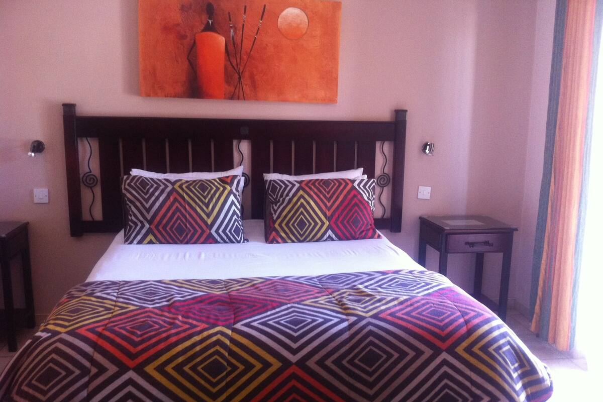 A bed with a patterned bedspread and two pillows, a nightstand, a wall-mounted artwork, and a window with curtains.