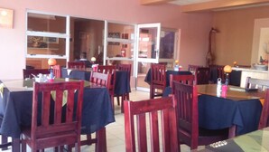 Un restaurant amb diverses taules i caires, una zona de cuina visible a través de portes de vidre, i un extintor fixat a la paret.