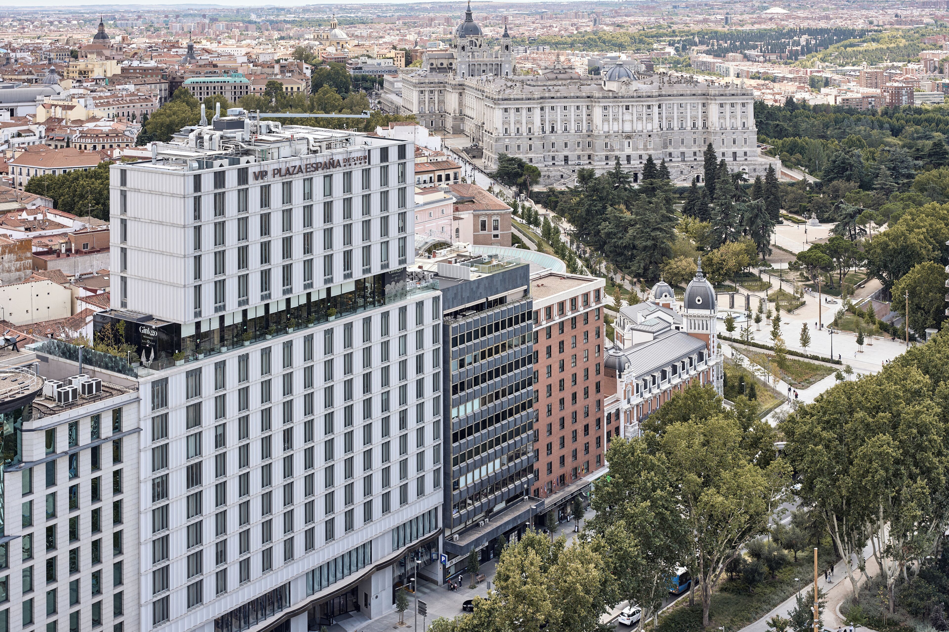 A modern hotel with a prominent glass facade, surrounded by historic buildings and lush greenery.