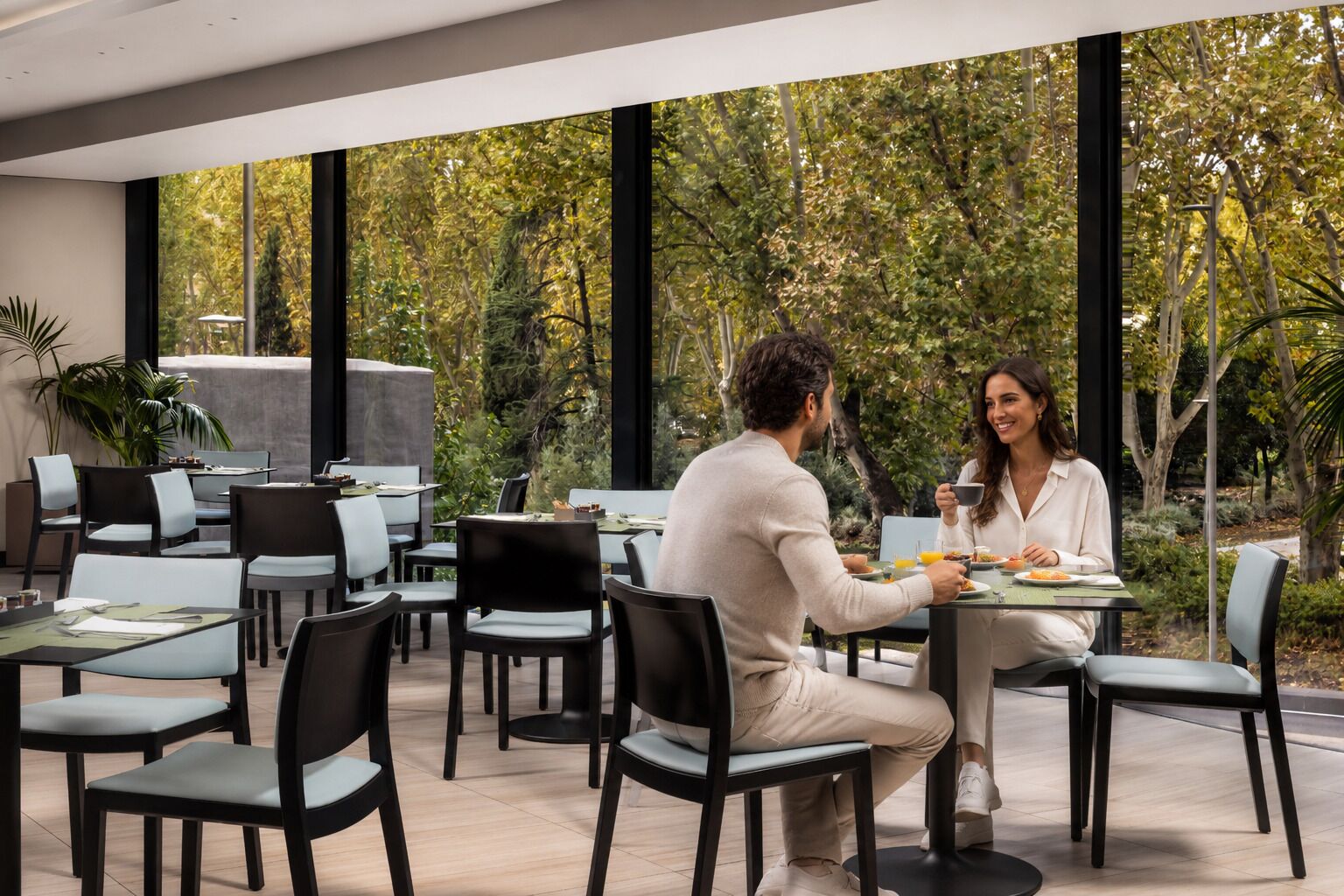 A modern outdoor dining area with tables and chairs, surrounded by greenery and large windows.