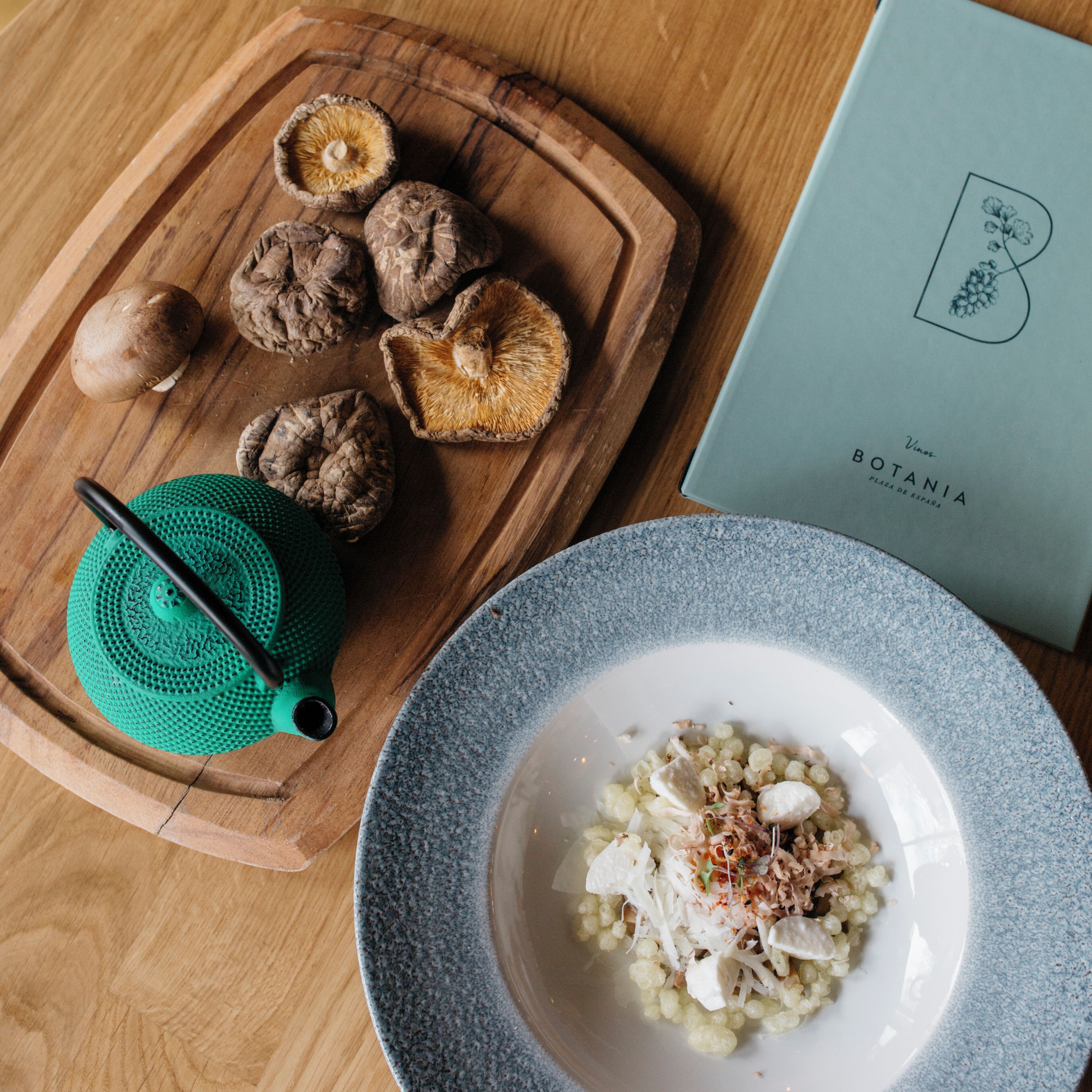 Un tagliere di legno con funghi e un colino verde, un piatto con un vassoio di cibo, e un libro intitolato "Vinos Botania".