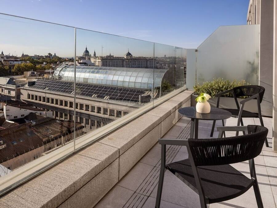 Un balcón con barandilla de cristal que ofrece vistas a un paisaje urbano, incluyendo un gran edificio de cristal y arquitectura tradicional.
