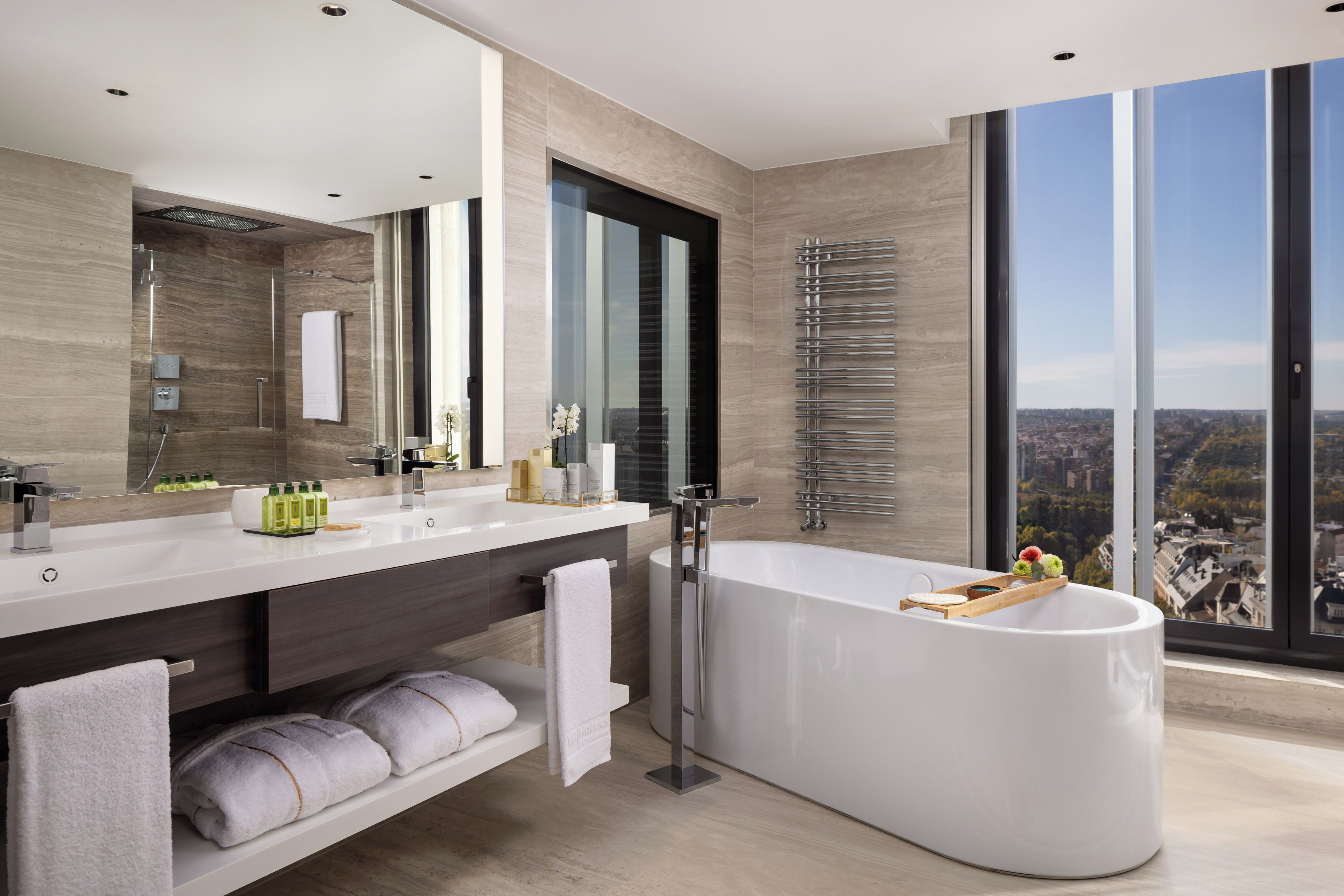 A modern bathroom with a large bathtub, a vanity with a mirror, and a view of the city through large windows.
