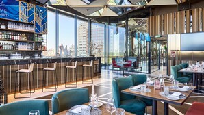 Un restaurante moderno con zona de bar, mesas y sillas, y grandes ventanales con vistas a la ciudad.