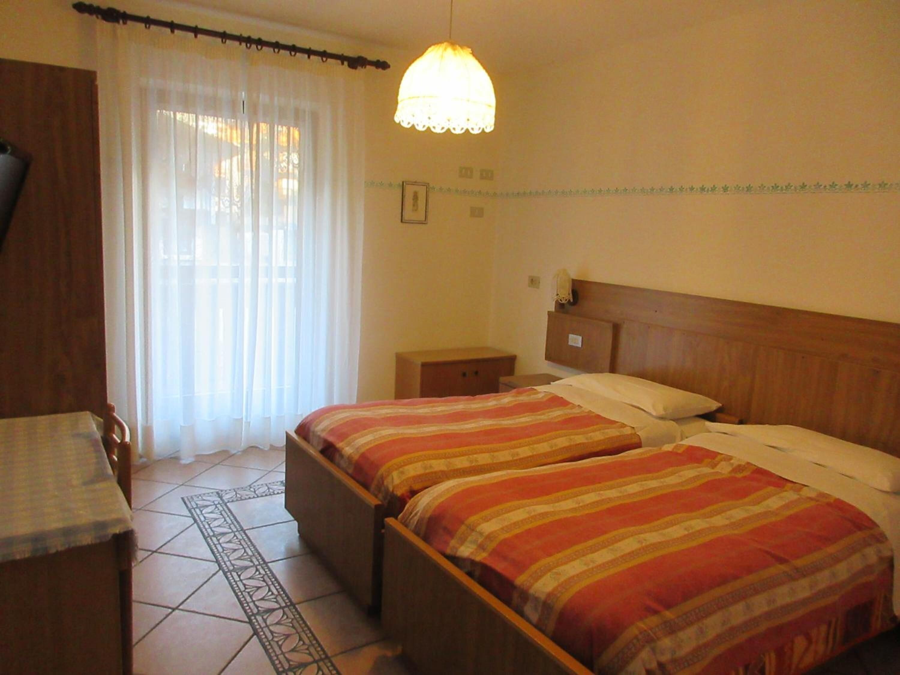 Habitación de hotel con dos camas, cabecera de madera, mesita de noche, lámpara y ventana con cortinas traslúcidas.