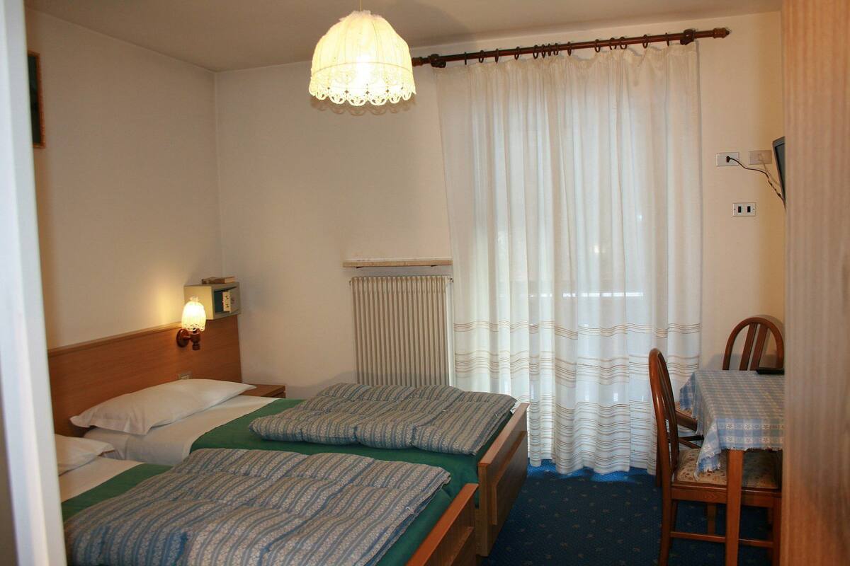 A hotel room with two beds, a wooden headboard, a table with a tablecloth, a chair, a hanging light fixture, and a window with curtains.