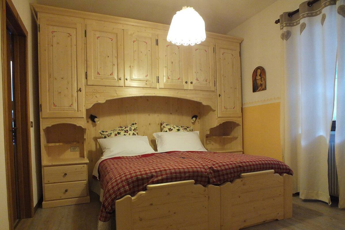 A wooden bed with a red checkered bedspread, two pillows, and a nightstand. There are wooden cabinets above the bed and a small nightstand to the left.