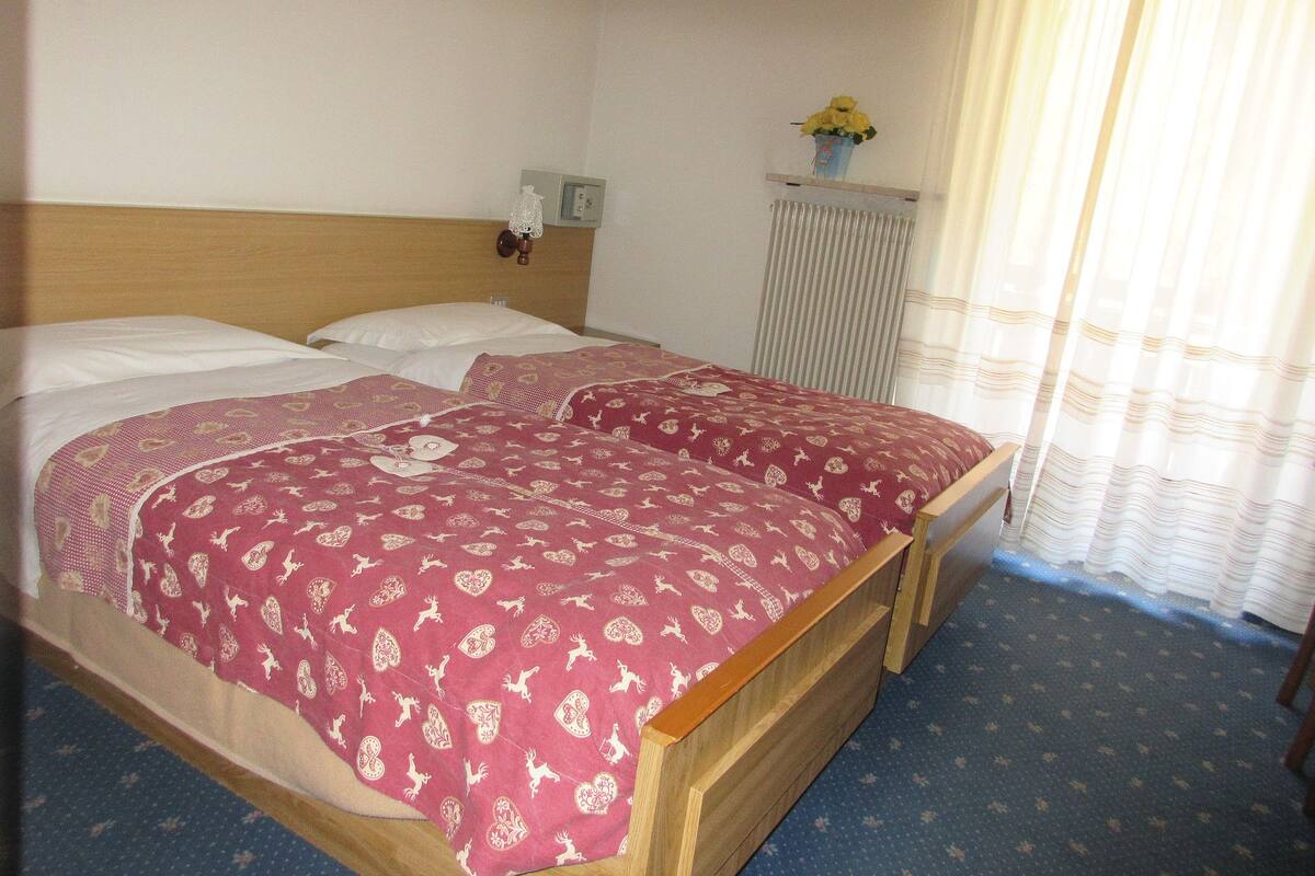 A double bed with patterned bedding, a wooden headboard, a bedside table with a lamp, a radiator, and a window with curtains.