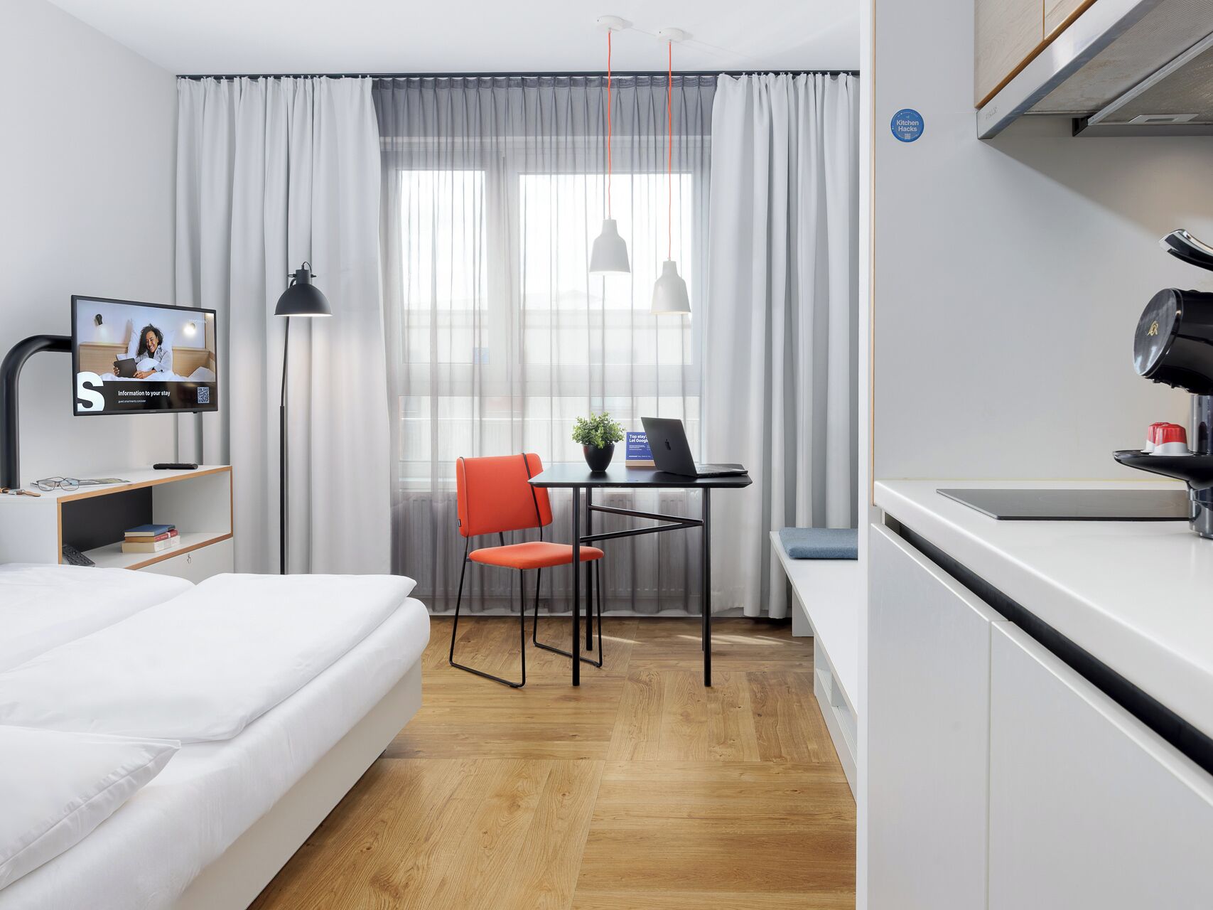 A modern hotel room with a bed, a desk with a laptop, a chair, a television, and a kitchenette.