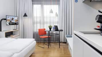 A modern hotel room with a bed, a desk with a laptop, a chair, a television, and a kitchenette.