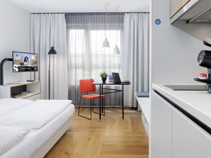 A modern hotel room with a bed, a desk with a laptop, a chair, a television, and a kitchenette.
