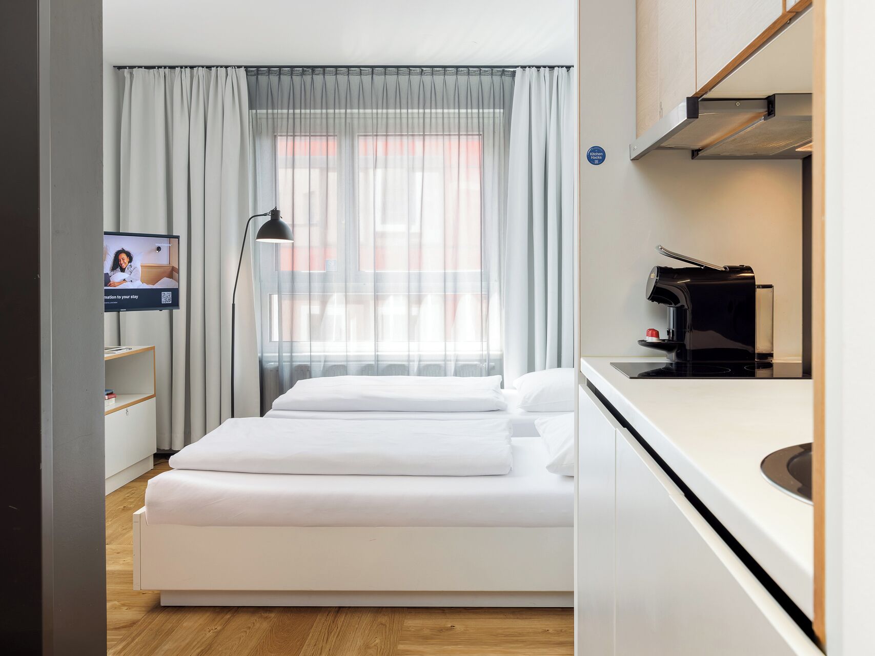 A hotel room with a bed, a desk with a coffee machine, a TV on the wall, and a window with curtains.
