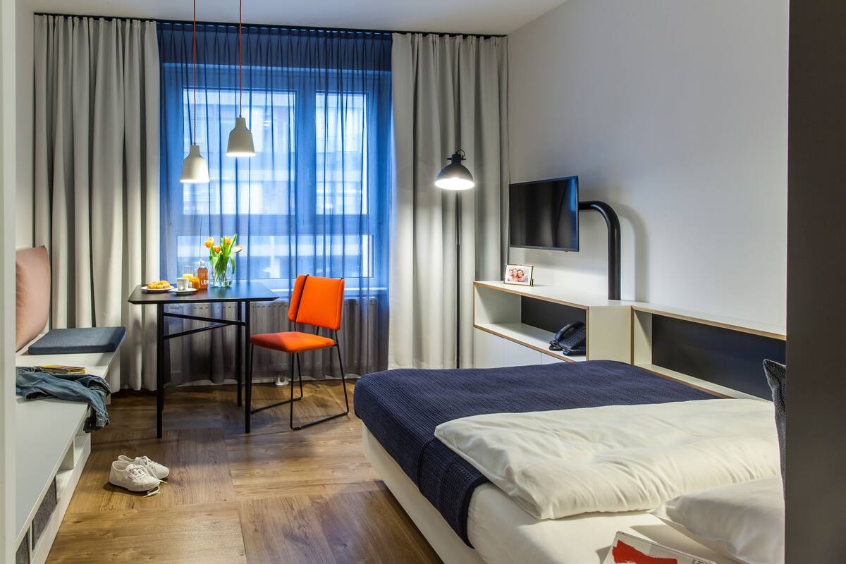 Une chambre d’hôtel avec un lit, un bureau, une chaise, une télévision et une fenêtre avec des rideaux.