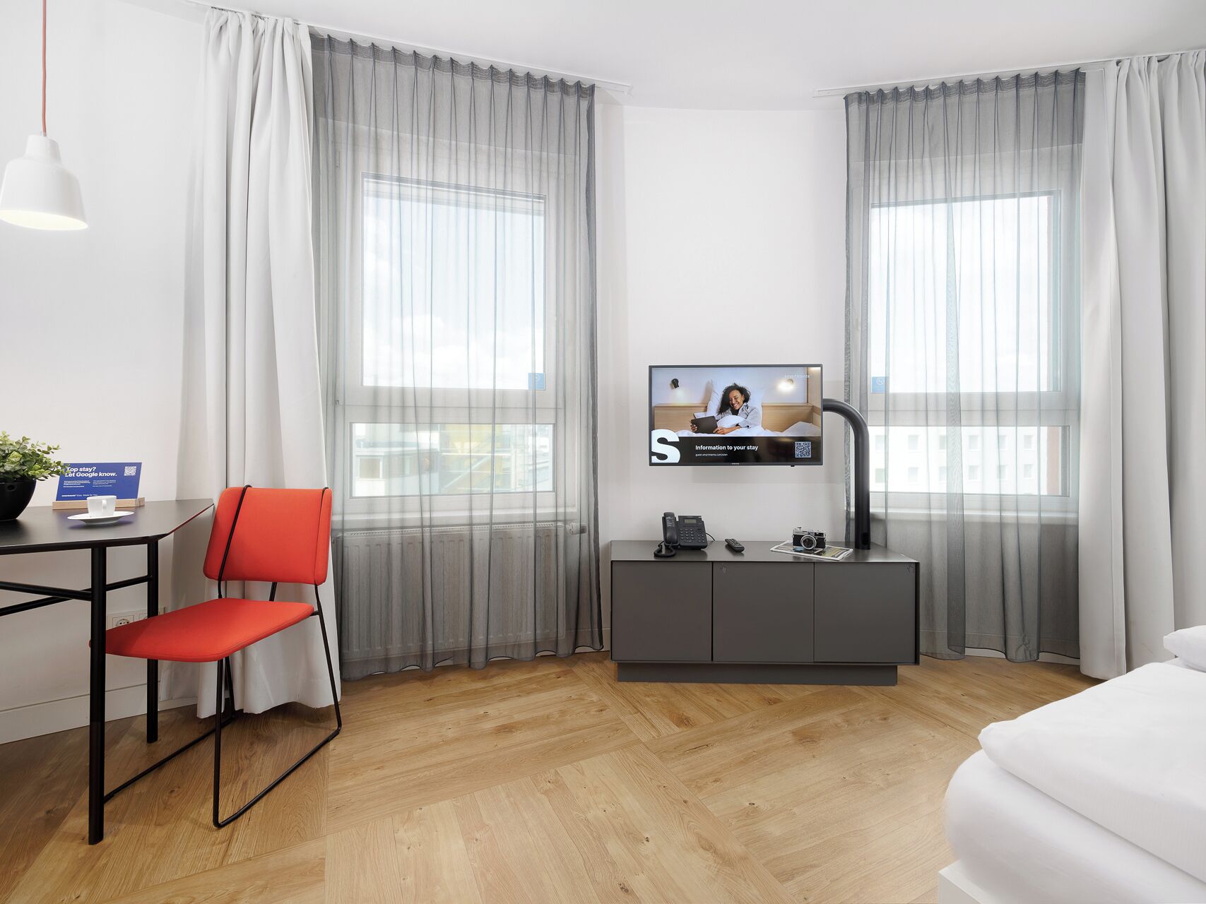 A hotel room with a bed, a desk with a chair, a television, and a large window with curtains.