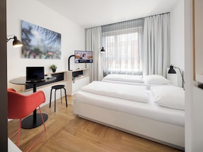 Une chambre d’hôtel moderne, équipée d’un lit, d’un bureau, d’une chaise, d’une télévision et offrant une vue sur la ville à travers de fines rideaux.