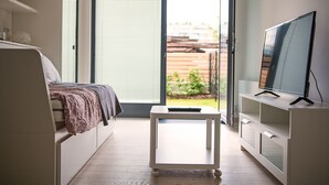 A modern bedroom with a bed, a small table with a remote control, a TV on a stand, and a sliding glass door leading to a garden. - Trinity Residence (Prague)