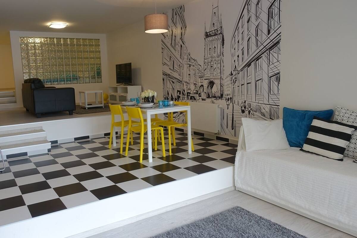 Un salon avec un sol à carreaux, un canapé blanc agrémenté de coussins bleus et noirs, une table à manger avec des chaises jaunes et une grande fresque murale.