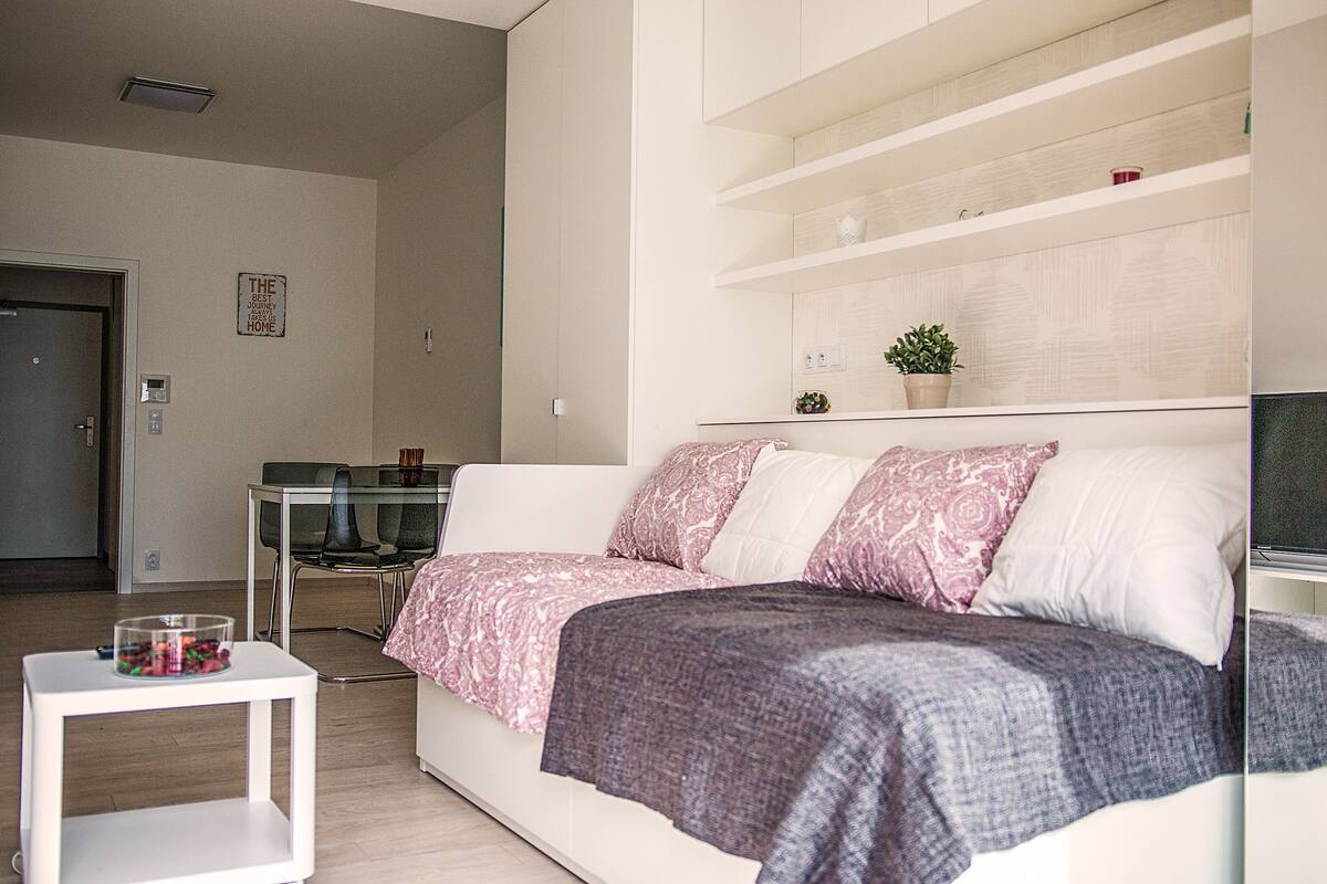 Une chambre à coucher moderne avec un canapé blanc, une petite table blanche surmontée d’un récipient en verre et une étagère intégrée agrémentée d’objets décoratifs.