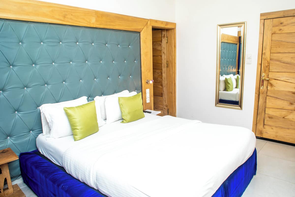 Une chambre à coucher avec une tête de lit capitonnée, un lit recouvert d’une literie blanche et verte, une table de chevet en bois et un miroir accroché au mur.