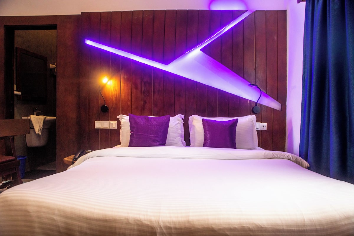 Une chambre d’hôtel avec un lit, des murs recouverts de panneaux en bois, un luminaire fixé au mur et une salle de bain visible au fond.