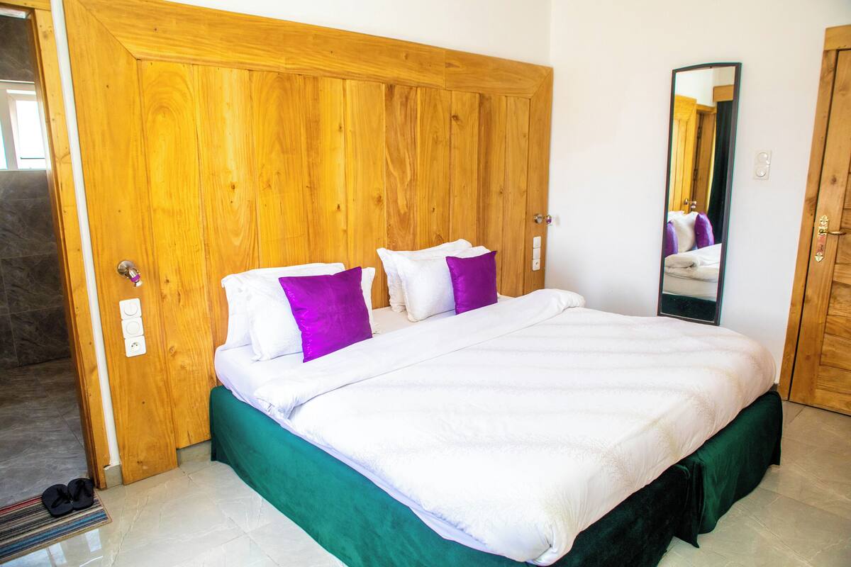 Une chambre avec une armoire en bois, un lit avec une literie blanche et violette, et un miroir au mur.