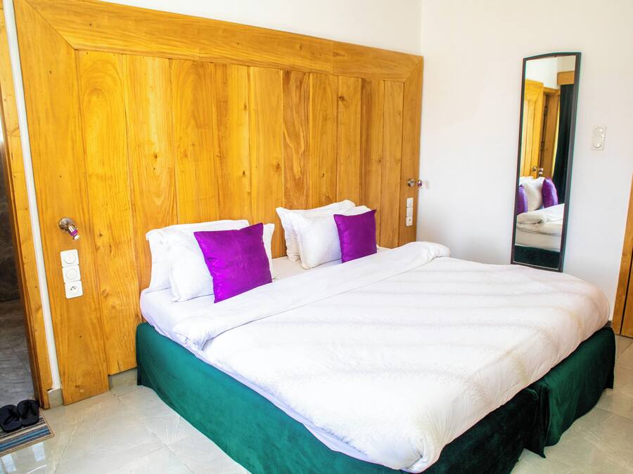 Un dormitorio con un armario de madera, una cama con ropa de cama blanca y morada, y un espejo en la pared.