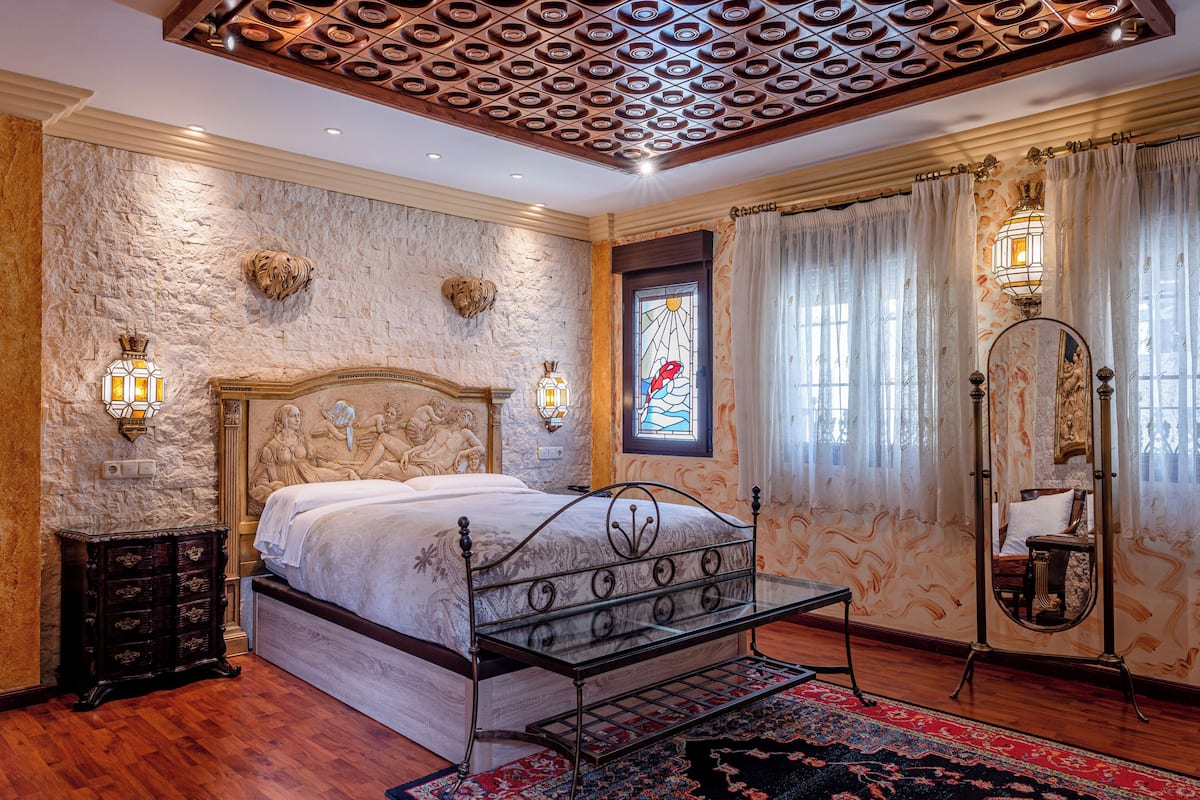Un dormitorio con pared de piedra, una cama con estructura metálica, un aparador de madera, un espejo y una ventana con vitrales.