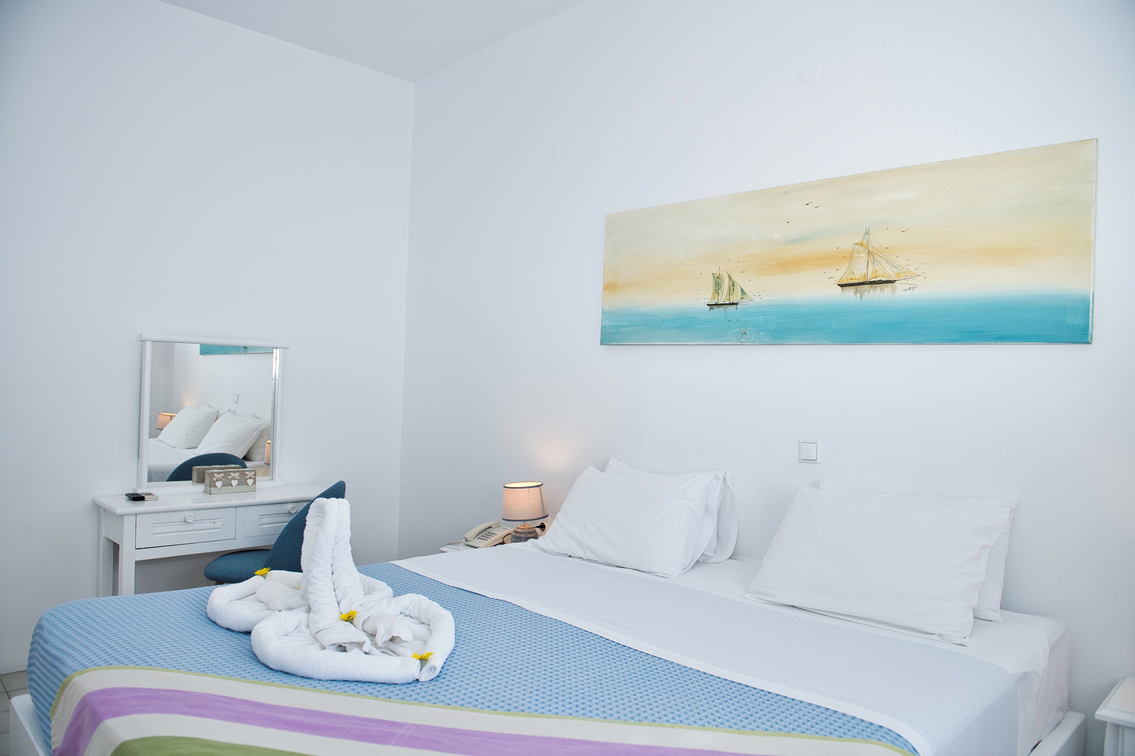 A hotel room with a bed, a desk, a chair, a mirror, a painting of a sailboat, and a lamp.