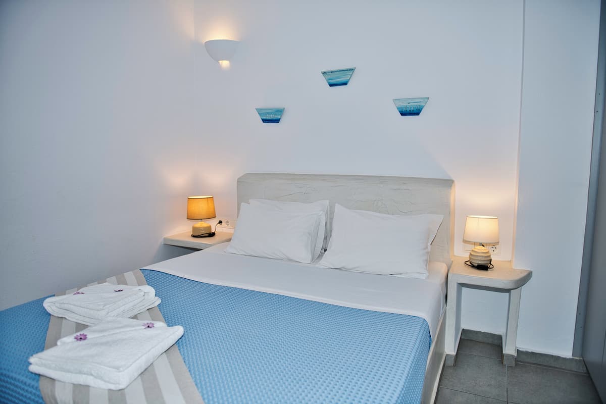 Una cama bien hecha con sábanas blancas y un almohadón a rayas azules, dos lámparas de noche y un aplique en la pared.